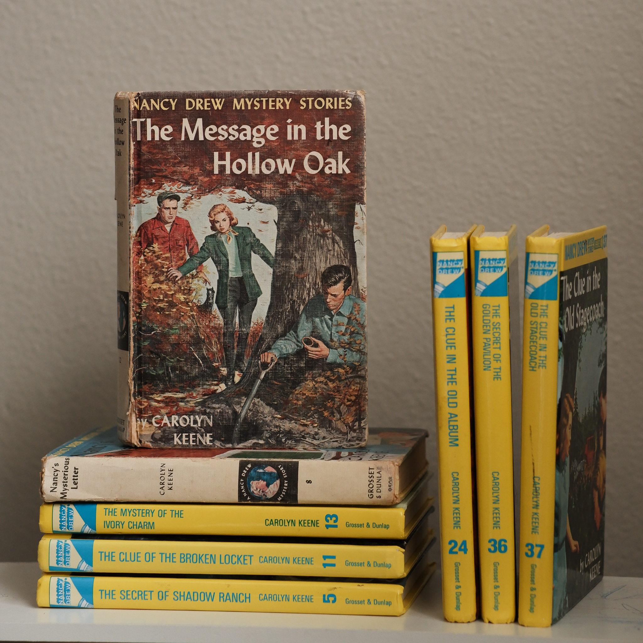 A collection of vintage Nancy Drew books, with the cover of The Message in the Hollow Oak showing. The cover shows a strawberry blonde young woman walking through the trees, and two men that she's probably about to investigate.