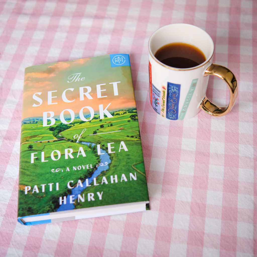 Hardcover Book of the Month Club edition of The Secret Book of Flora Lea, a novel by Patti Callahan Henry. Pictured on a pink checked tablecloth next to a cup of coffee.