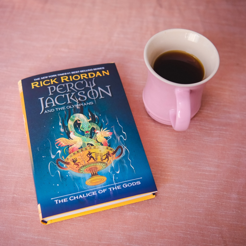 A hardcover copy of The Chalice of the Gods by Rick Riordan, sitting next to a pink mug filled with coffee.