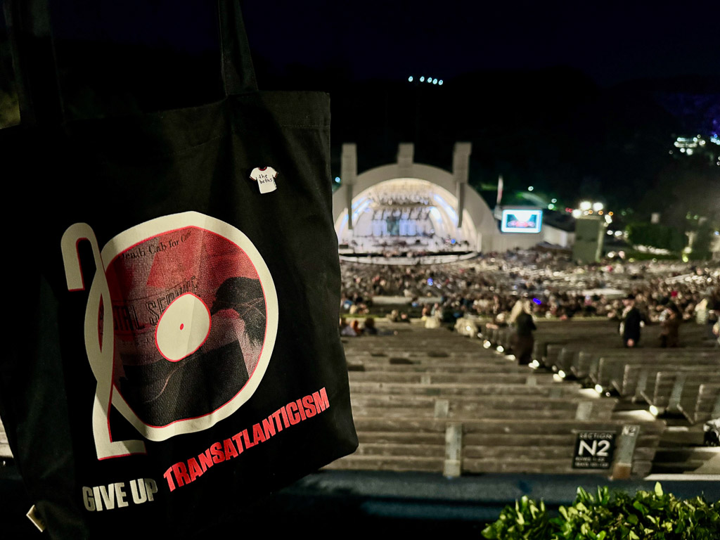 Black tote bag with the logo for the Death Cab for Cutie and The Postal Service 20th anniversary tour for Give Up and Transatlanticism. The tote bag has an added pin that looks like a white t-shirt which says The Beths on it. The Hollywood Bowl can be seen in the background.