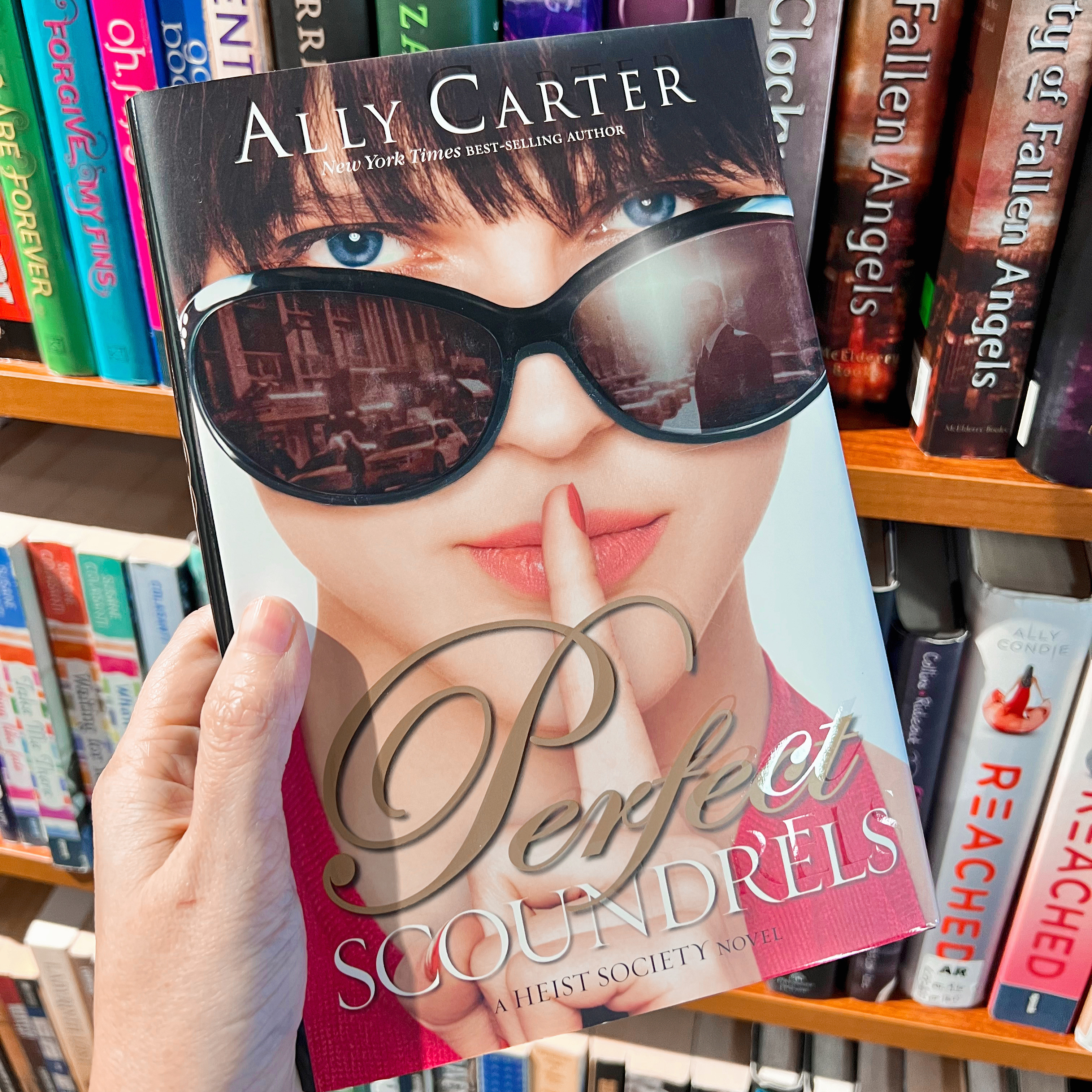 A hand holding a book with books on shelves behind it. The book is Perfect Scoundrels by Ally Carter.