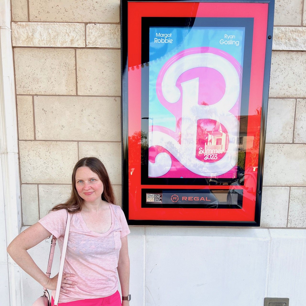 Shannon standing in front of the poster for the Barbie movie. Shannon is wearing a light pink shirt with dark pink shorts.