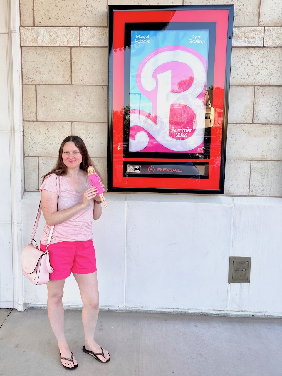Shannon standing in front of the poster for the Barbie movie. Shannon is wearing a light pink shirt with dark pink shorts. Shannon is holding Barbie doll as Elle Woods from Legally Blonde 2.